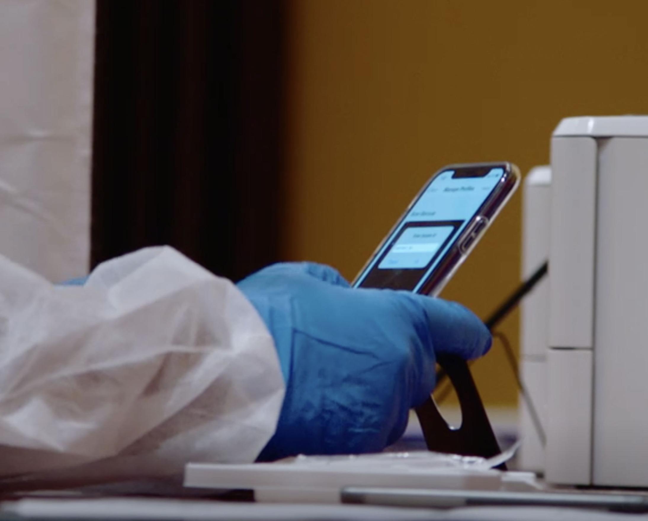 A gloved hand from a person in a lab coat holds a smartphone